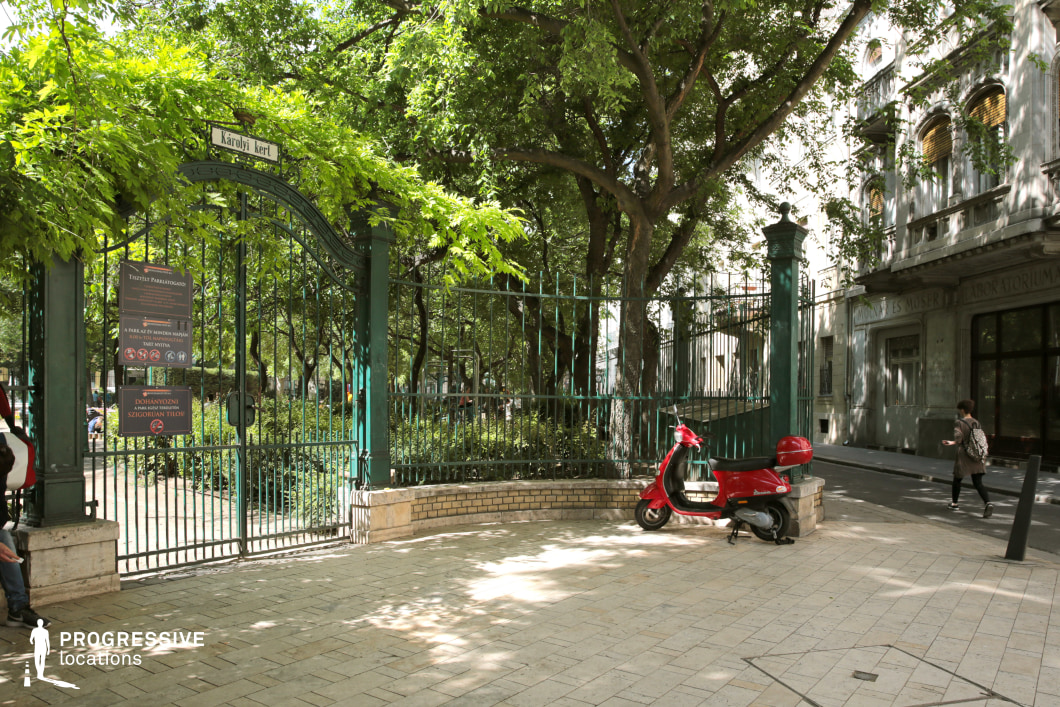 A wrought-iron gate with intricate detailing marks the entrance to a lush urban park, framed by tall, mature trees that create a natural canopy. The gate's dark green color complements the surrounding vegetation, while a low brick wall and stone pavement define the transition between the park and the street. Adjacent to the entrance, a historic stone building with ornate balconies and arched windows adds an old-world charm to the scene. The soft contrast between the greenery and the structured urban elements makes this location visually compelling. This setting is ideal for filming scenes requiring a refined yet accessible public space, suitable for genres such as drama, romance, and historical narratives.