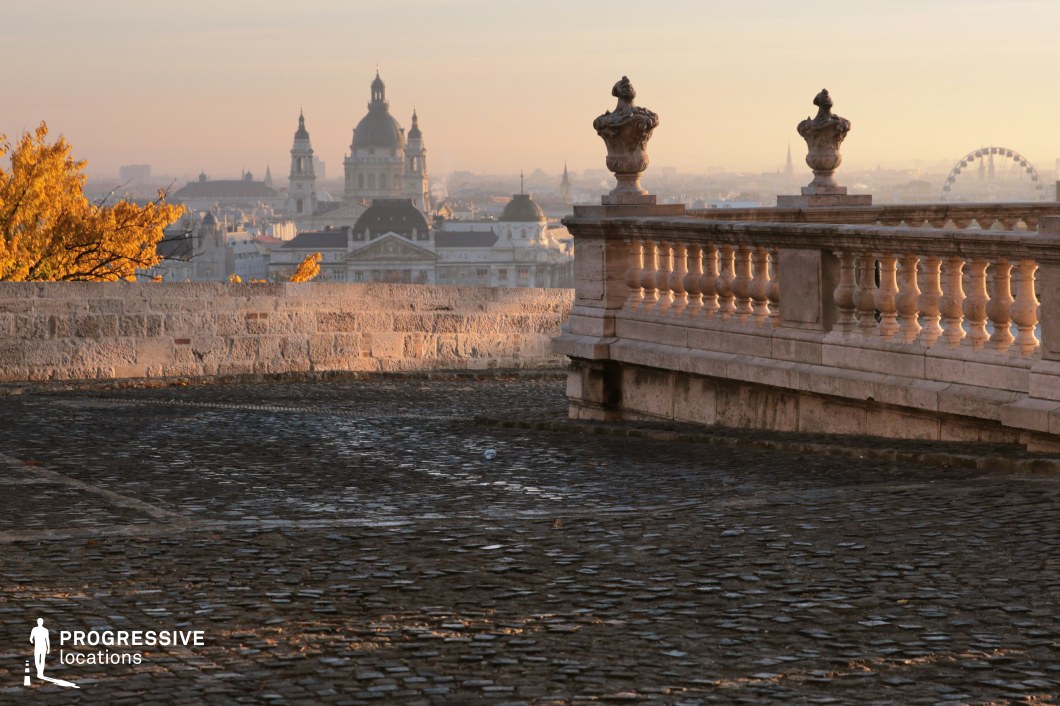 This image captures a historic European terrace with an intricately carved stone balustrade and classical urn-shaped ornaments overlooking a grand cityscape. The textured cobblestone pavement leads towards a distant skyline featuring domed landmarks, elegant rooftops, and a Ferris wheel, creating a rich visual depth. A weathered stone wall encloses part of the terrace, reinforcing the location’s historic charm. This setting is ideal for filming period dramas, reflective moments, or grand city introductions, offering a cinematic backdrop that evokes timeless sophistication and old-world elegance.
