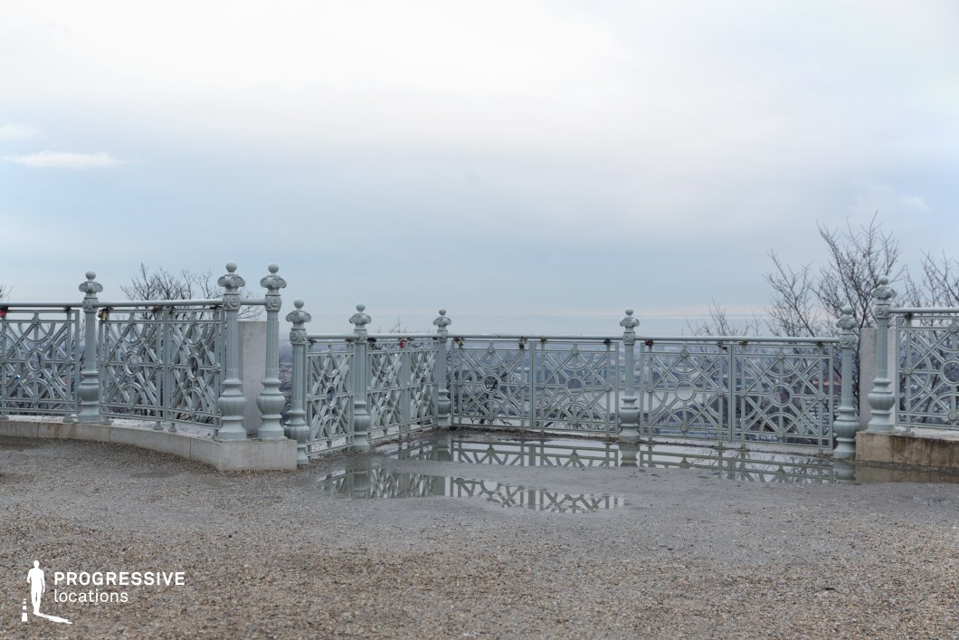 A scenic viewpoint enclosed by an intricately designed wrought-iron railing, featuring symmetrical patterns and decorative finials that enhance its refined, classical appearance. The gravel ground, slightly damp, reflects the ornate railing, adding depth to the composition. The open space offers an expansive view of the cityscape below, framed by bare trees that emphasize the location’s elevated perspective. This setting is ideal for contemplative, dramatic, or romantic scenes, serving as a visually striking backdrop for period films, historical narratives, or emotionally charged storytelling.