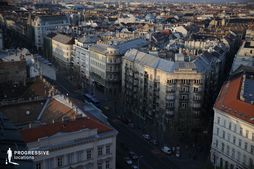 A panoramic view of a dense urban landscape showcasing a mix of historic and modern buildings, characterized by intricate facades, sloping rooftops, and varied architectural styles. The streets below weave through the tightly packed structures, with intersections creating natural focal points. The curved corner buildings and metal rooftops add a distinctive European character, while distant domes and taller structures provide depth and scale. This location is ideal for aerial establishing shots in films, providing a visually rich setting for historical dramas, crime thrillers, or atmospheric city-based storytelling.