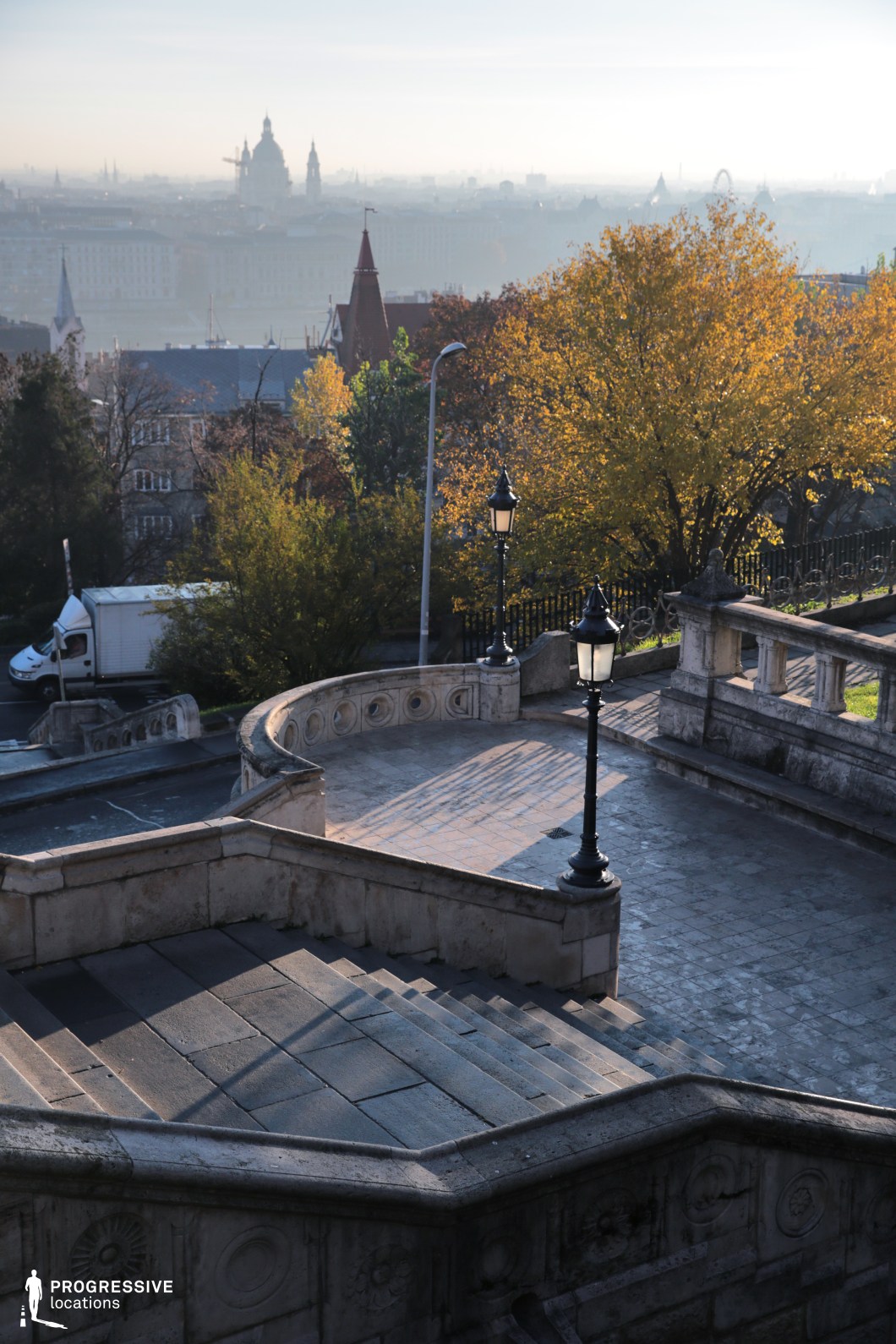 This image captures a grand stone staircase descending into a European cityscape, framed by ornate balustrades and elegant street lamps. The curved landings and intricate circular relief details on the stone walls add a touch of historic refinement. Beyond the staircase, the view opens to a sweeping city skyline featuring architectural landmarks, rooftops, and church domes, creating a sense of depth and timeless grandeur. The scene is rich in cinematic potential, ideal for historical dramas, romantic encounters, or contemplative urban sequences, offering a visually layered and atmospheric setting reminiscent of classic European cities.
