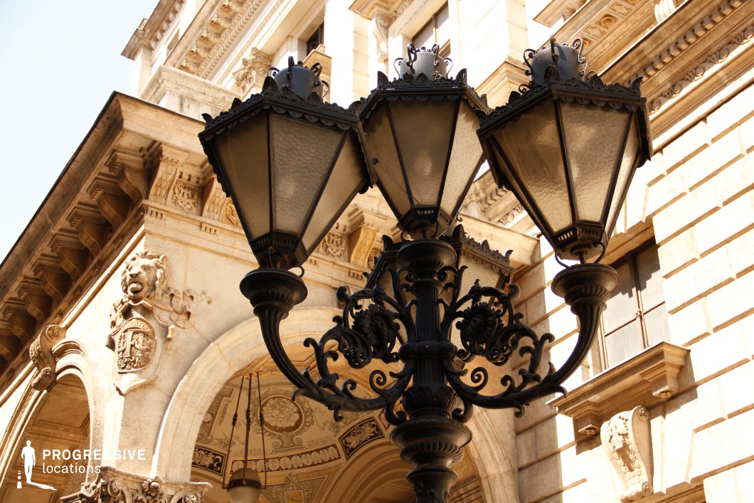 The image highlights an intricately designed black wrought-iron street lamp with three lanterns, adorned with elaborate scrollwork and decorative finials. The background features a historic stone building with grand archways, sculpted reliefs, and detailed cornices, reinforcing the classical European aesthetic. A lion crest and ornamental carvings add further elegance to the architecture. The perspective emphasizes the lamp’s intricate craftsmanship while integrating it seamlessly into the surrounding historic environment. This location is ideal for filming period dramas, aristocratic settings, or mystery narratives, offering a visually rich and sophisticated old-world atmosphere.