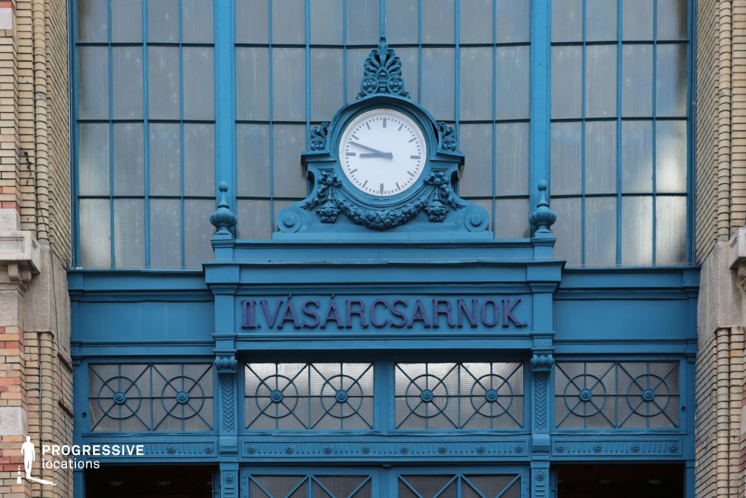 A striking blue entrance of a historic market hall, featuring intricate ironwork, large glass panels, and a central clock framed by elaborate sculptural details. The deep blue color contrasts with the surrounding brick and stone elements, highlighting the building’s industrial-era elegance. Decorative finials and geometric latticework on the doors add to the architectural richness, while the large glass windows allow natural light to filter through. This location is ideal for filming scenes requiring a historic yet functional urban setting, making it suitable for period dramas, European cityscapes, or atmospheric mystery and noir genres.