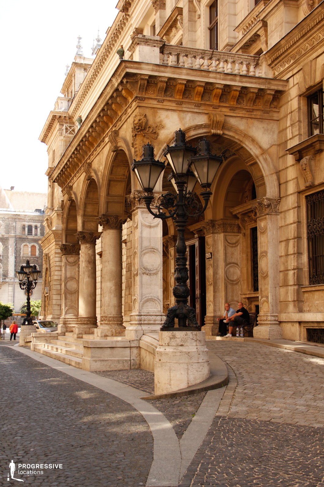 This image captures a striking neoclassical or baroque-style building with intricate architectural details, including grand arched entrances, sculpted reliefs, and decorative cornices. The elegant stone façade features a weathered yet sophisticated appearance, enhanced by massive columns supporting a richly adorned portico. The curved cobblestone street in the foreground adds depth and a sense of movement, while the ornate black cast-iron street lamps reinforce the historic European atmosphere. This location is ideal for filming period dramas, historical films, and sophisticated cityscapes, offering an authentic old-world charm reminiscent of classical European cities.