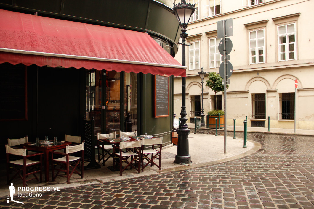 This image captures a classic European café situated on a street corner, featuring a deep green façade with a contrasting red awning that extends over the neatly arranged outdoor seating. The dark wooden tables and canvas-backed chairs add a rustic charm, while the cobblestone street curves gently around the café, enhancing the old-world aesthetic. Ornate black street lamps and historic cream-colored buildings with arched windows contribute to the authentic European ambiance. This location is ideal for filming romantic dramas, intimate conversations, or atmospheric city scenes, offering a timeless and picturesque backdrop evocative of Parisian or old-world European settings.