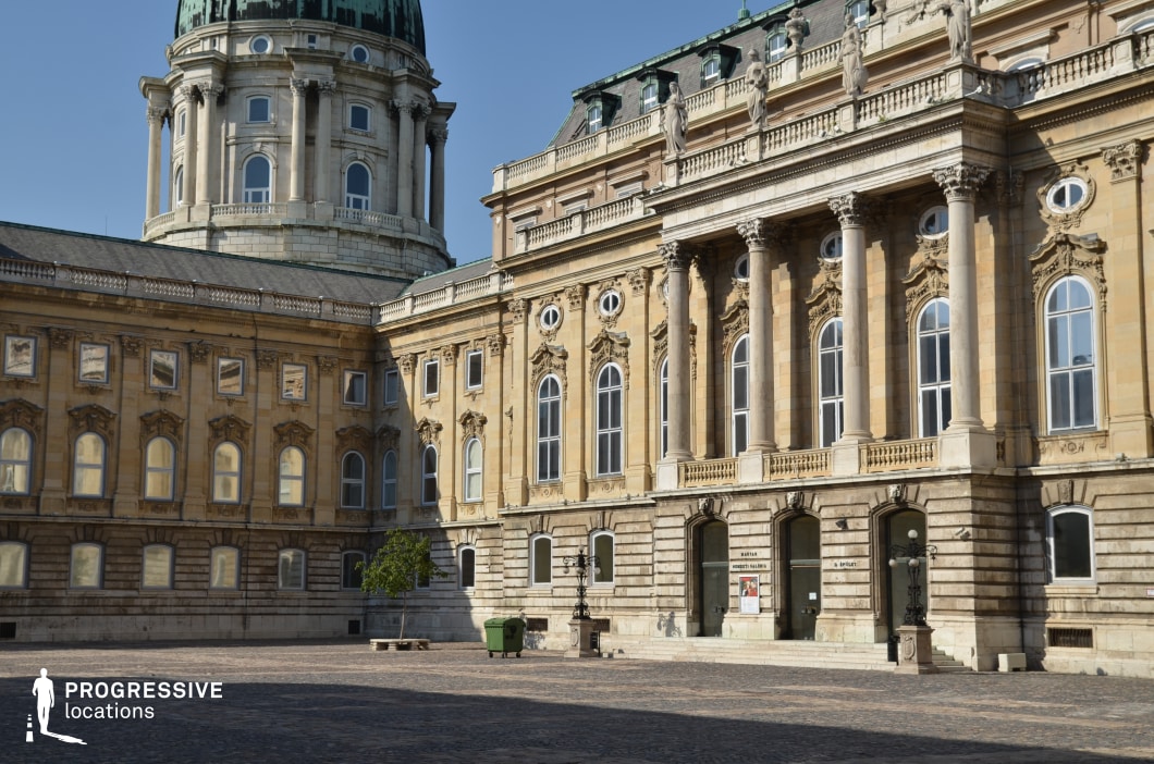 The image captures a grand courtyard enclosed by a historic palace featuring intricate Baroque and Neoclassical architectural details. Towering columns, sculpted reliefs, and decorative statues line the building’s facade, reinforcing its opulent and regal aesthetic. The large dome in the background, with its patinated green roof, adds a sense of monumentality to the scene. The cobblestone ground enhances the historical atmosphere, creating a timeless setting. This location is ideal for filming period dramas, royal narratives, or historical action films, providing a majestic and authentic European backdrop that exudes grandeur and sophistication.