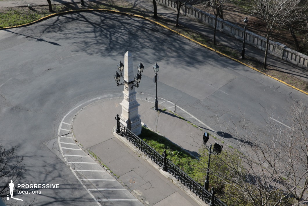 The image captures a small triangular traffic island featuring an elegant white stone obelisk adorned with intricate black wrought-iron street lamps. A decorative metal fence surrounds the monument, enhancing its historic character. The setting is framed by a curving road with aged asphalt and a raised stone walkway lined with balustrades, giving a sense of old-world charm. Shadows from nearby trees add depth and texture to the scene. This location is ideal for filming period pieces, urban dramas, or noir-inspired thrillers, offering a visually striking intersection that blends classical elegance with a slightly weathered atmosphere.