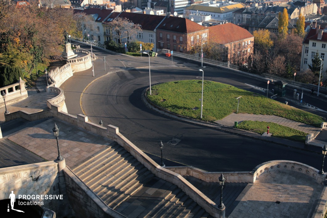 This image captures an impressive stone staircase leading down toward a curved road encircling a small green space, with a mix of historic and modern buildings in the background. The staircase features ornate balustrades and vintage-style street lamps, reinforcing its classic European character. The winding road adds a dynamic visual element, while the surrounding architecture, with red-tiled roofs and varied facades, creates a rich urban backdrop. This location is ideal for filming historical dramas, action sequences, or atmospheric cityscape scenes, offering depth, movement, and a blend of elegance with urban functionality.