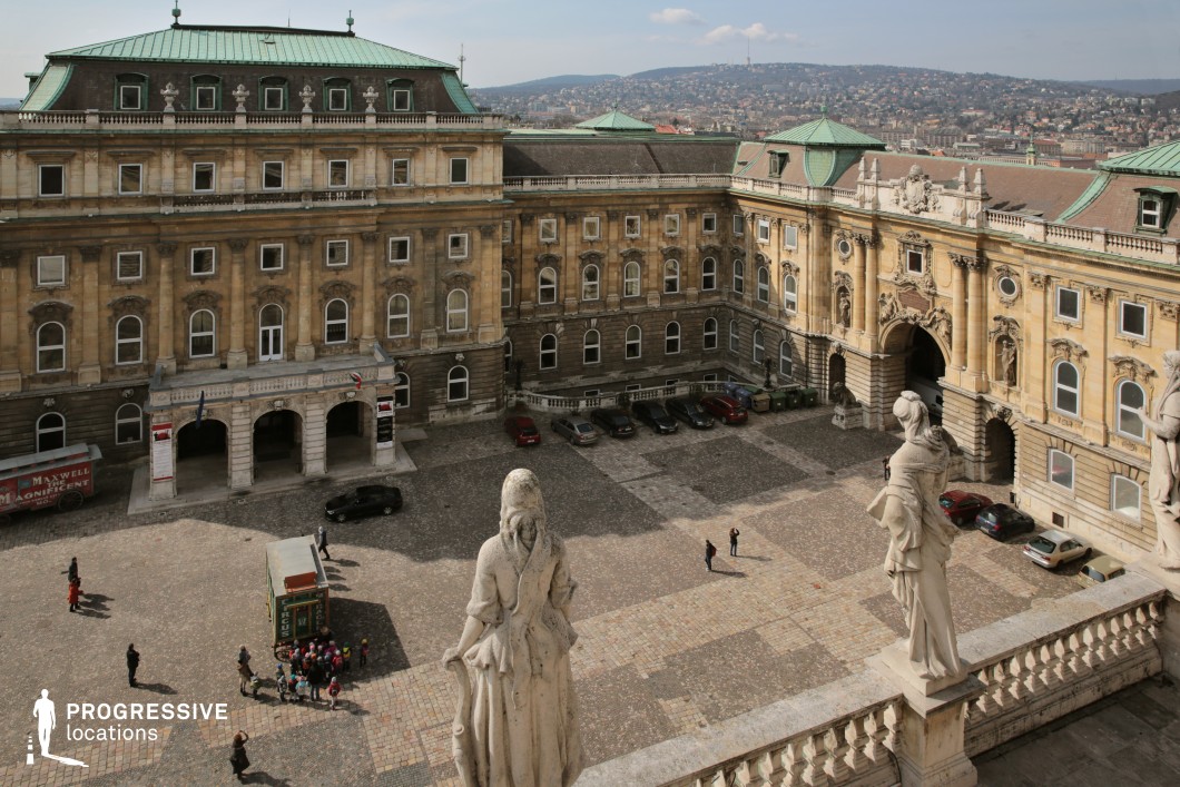 The image showcases a grand castle courtyard enclosed by historic buildings featuring intricate stonework, arched windows, and green patina-covered rooftops. The central arched gateway is adorned with elaborate carvings and statues, reinforcing the regal and classical aesthetic. The cobblestone courtyard is framed by a decorative balustrade with weathered stone statues overlooking the scene, adding a sense of historical depth. The backdrop reveals a sprawling cityscape with rolling hills, enhancing the sense of grandeur and openness. This location is ideal for filming historical dramas, period pieces, and regal intrigue, offering an authentic and visually captivating setting for aristocratic, royal, or medieval-themed productions.