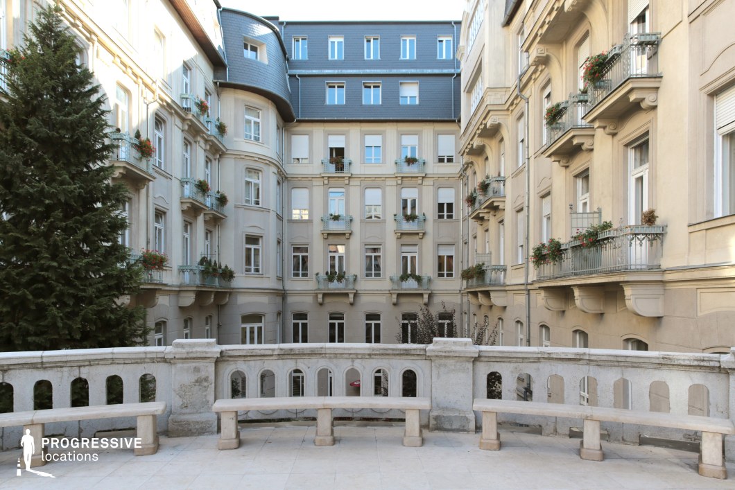 This image captures a beautifully enclosed European courtyard framed by classic beige-hued buildings with intricate architectural details. The façades feature decorative moldings, arched windows, and wrought-iron balconies adorned with flower arrangements, creating a refined and harmonious aesthetic. A stone balustrade with built-in benches overlooks the space, providing a serene focal point. The upper floors display a mansard-style roof with dormer windows, reinforcing the historic charm of the setting. This location is ideal for filming period dramas, romantic films, or sophisticated urban scenes, offering an intimate and timeless European atmosphere.