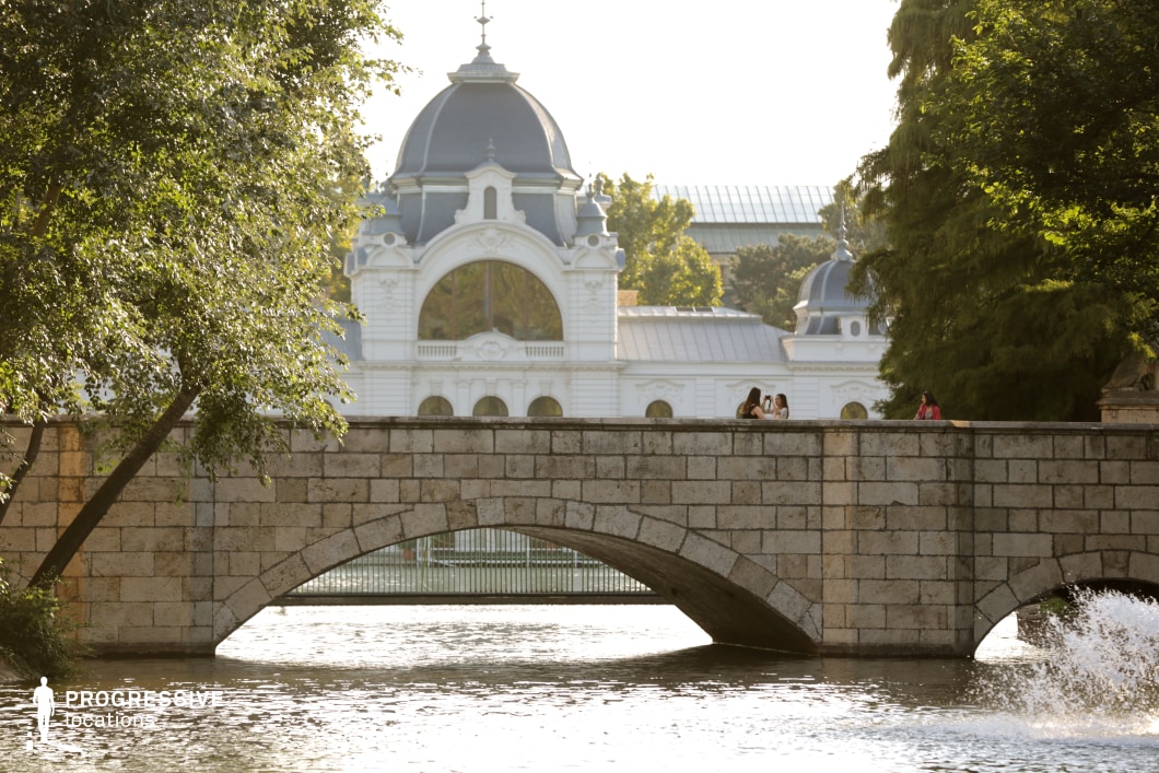 This image features a picturesque stone bridge with elegant archways spanning a reflective body of water. The bridge, built from large, textured stone blocks, exudes a sense of timeless solidity. In the background, a grand neoclassical building with ornate white facades and domed rooftops adds a striking visual contrast, framed by lush green trees. The scene is rich in architectural harmony, with the bridge and the building forming a balanced composition. This location is ideal for filming romantic, historical, or atmospheric drama scenes, offering a blend of European elegance and scenic tranquility that enhances both period and contemporary storytelling.