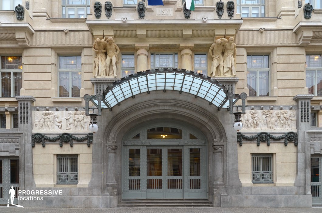 The image showcases a striking Art Nouveau entrance framed by an elegant arched doorway with intricate metalwork and frosted glass panels. Above the entrance, a curved glass canopy with decorative iron supports enhances the building’s refined aesthetic. The facade features detailed bas-reliefs depicting classical scenes and two imposing statues of muscular figures flanking the upper level, reinforcing the monumental character of the structure. Ornate street lamps and sculptural embellishments further emphasize the historic and artistic significance of the location. This setting is ideal for filming period dramas, high-society intrigue, or elegant urban sequences, offering a richly detailed and architecturally impressive backdrop.