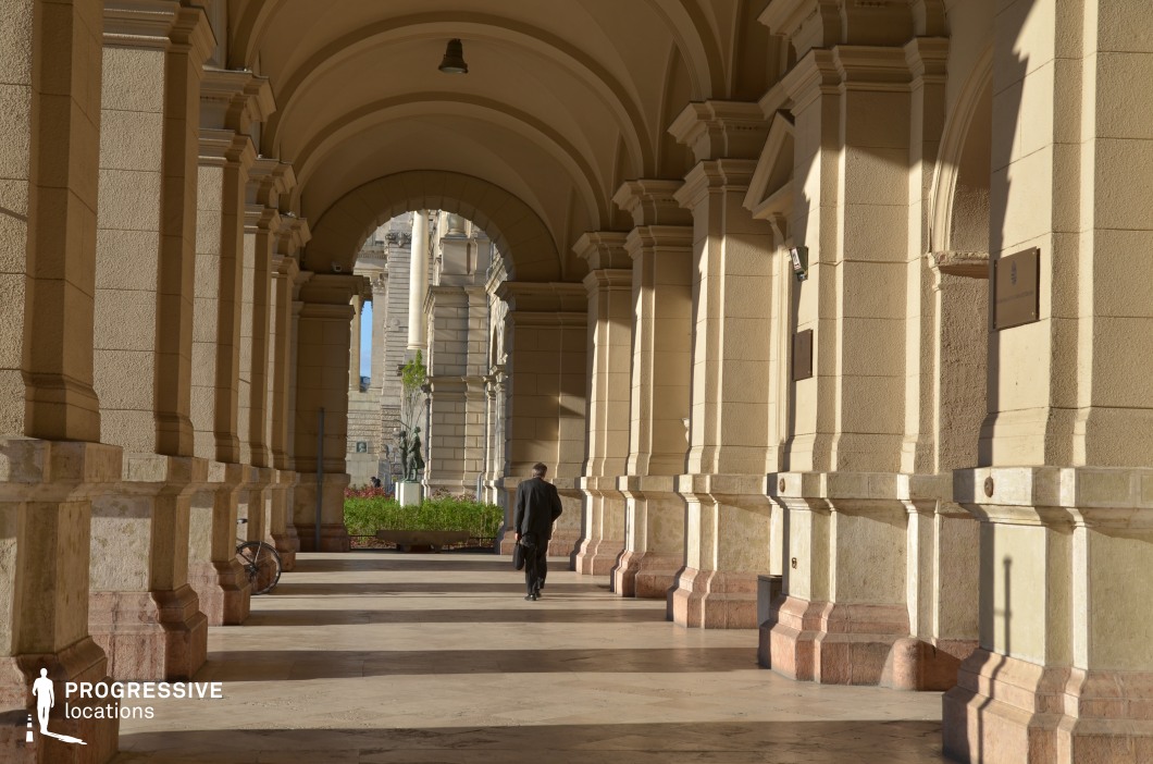 The image showcases an elegant covered walkway lined with massive classical columns supporting a series of symmetrical arches. The soft beige and cream-colored stone contrasts with the polished marble floor, while the depth created by repeating archways adds a dramatic visual effect. Natural light filters through, casting defined shadows that emphasize the architectural rhythm. The passage leads towards an open courtyard framed by historic buildings, enhancing the sense of grandeur. This location is ideal for filming historical dramas, political thrillers, or sophisticated urban narratives, offering a refined, neoclassical setting with strong cinematic depth.