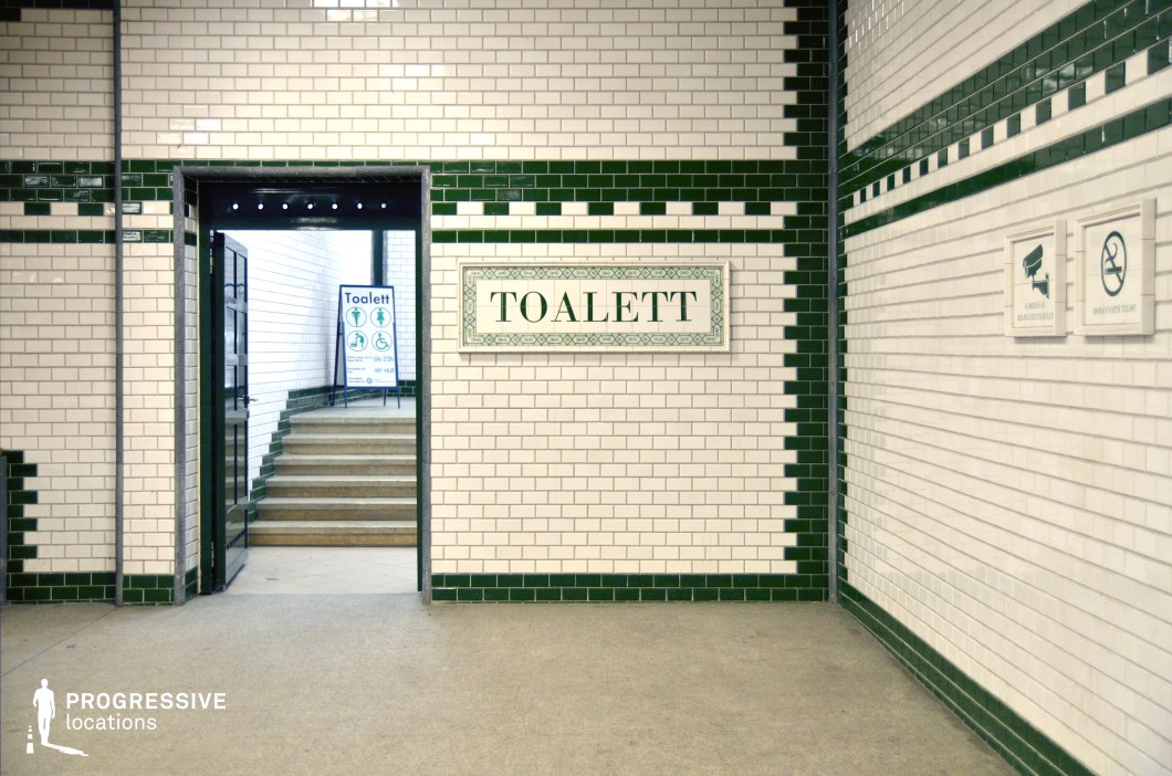 A clean, tiled underground corridor with a striking vintage aesthetic, featuring white subway tiles accented by deep green borders in a symmetrical pattern. The bold typography on the decorative sign and the classic mosaic-style framing contribute to the historical charm of the space. The open doorway leads to a softly lit stairway, creating a sense of depth and perspective. The polished floor reflects ambient light, enhancing the sterile yet structured atmosphere. This location is ideal for filming suspenseful, noir, or period scenes, offering a timeless setting reminiscent of early 20th-century urban infrastructure.