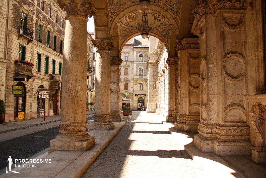 This image showcases a grand European archway supported by massive, intricately carved stone columns adorned with circular relief patterns and ornate capitals. The ceiling features elaborate decorative moldings and gilded embellishments, emphasizing the opulence of the structure. The perspective leads through the covered passageway toward a sunlit street framed by elegant historic buildings with tall windows and wrought-iron balconies. The combination of light and shadow adds depth, enhancing the architectural richness. This location is ideal for filming period dramas, historical epics, and sophisticated city scenes, offering a regal and timeless atmosphere reminiscent of classic European capitals.