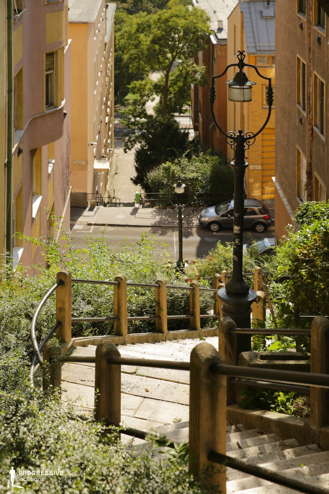 This image features a steep, winding urban stairway flanked by classic black wrought-iron lamp posts and bordered by weathered stone railings with metal handrails. The stairs descend into a picturesque narrow street lined with warm-toned residential buildings in shades of ochre, orange, and beige, creating a cozy, European atmosphere. Lush green foliage partially frames the stairway, softening the urban edges. The perspective emphasizes depth and movement, making this location ideal for dynamic chase scenes, contemplative character moments, or romantic urban storytelling. The charming old-world aesthetic lends itself well to dramas, thrillers, or nostalgic period films.