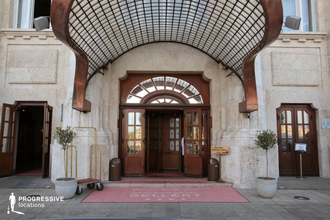 The image showcases the grand entrance of a historic hotel, featuring a distinctive curved metal and glass canopy in an Art Nouveau style. The entrance is framed by a symmetrical stone facade with wooden double doors and arched windows, emphasizing a refined, early 20th-century aesthetic. The elegant pink carpet leading inside enhances the luxurious feel, while the brass luggage cart and potted plants add to the welcoming atmosphere. This location is ideal for filming period dramas, sophisticated urban narratives, or vintage-inspired travel stories, offering an authentic and visually rich backdrop with classic European charm.