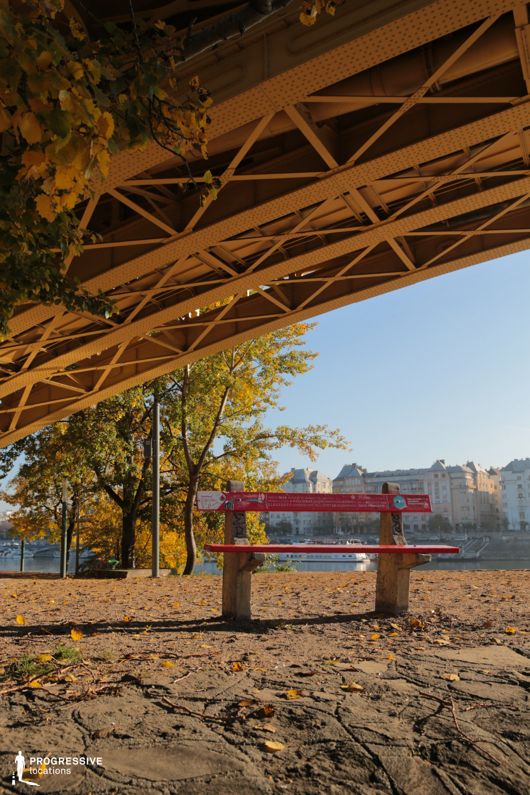 A sturdy metal bridge with intricate riveted beams stretches overhead, framing a peaceful riverside scene below. A red wooden bench stands on the textured ground, facing the calm river, offering an inviting resting spot with a panoramic view of classic European-style buildings in the distance. The combination of natural and industrial elements—leafy trees contrasting with the structured geometry of the bridge—creates a visually rich composition. This location is ideal for contemplative or introspective scenes, making it suitable for dramas, urban storytelling, or romantic narratives with a scenic city backdrop.