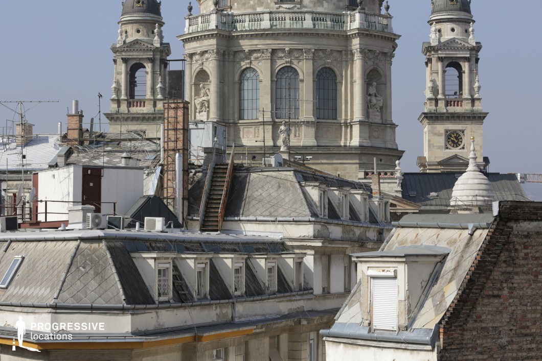This image presents a striking contrast between aged city rooftops and the grandeur of a historic basilica. The foreground consists of old mansard-style rooftops with weathered zinc panels, small dormer windows, and scattered urban elements such as ventilation systems and antennas, creating an authentic, lived-in cityscape. A steep, narrow staircase connects different rooftop levels, adding depth and an element of urban exploration. The background features an ornate basilica with towering bell towers, statues, and intricate stonework, reinforcing a sense of history and grandeur. This location is ideal for filming espionage thrillers, rooftop chases, or atmospheric European city scenes, offering a unique blend of urban grit and architectural elegance.