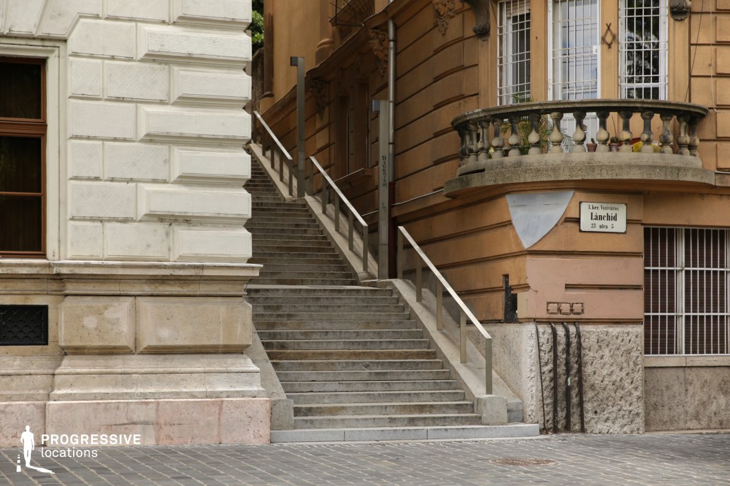 The image captures a steep stone staircase nestled between two historic buildings, leading to an upper level of the city. The architectural contrast between the light-colored, classical facade on the left and the warm-toned, ornate structure on the right adds visual depth. The right building features a curved balcony with decorative balustrades, enhancing the old-world charm. Metal railings line the stairs, while subtle urban details such as wall-mounted pipes and textured stone surfaces contribute to the scene’s character. This location is ideal for filming noir-inspired mysteries, chase sequences, or atmospheric period dramas, offering an intimate yet dynamic urban setting with strong vertical composition.