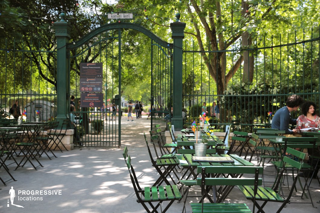 This image showcases a picturesque outdoor café set against the entrance of a lush, well-maintained park. The scene is framed by an elegant wrought-iron gate with decorative finials, flanked by sturdy green pillars. The café area features neatly arranged green metal tables and chairs, creating a relaxed, inviting atmosphere. A tree-lined path extends beyond the gate, leading into a verdant park filled with vibrant foliage and blooming flowers. The combination of natural and architectural elements makes this location ideal for filming romantic scenes, casual city life montages, or atmospheric daytime sequences, offering a serene and classic European setting.