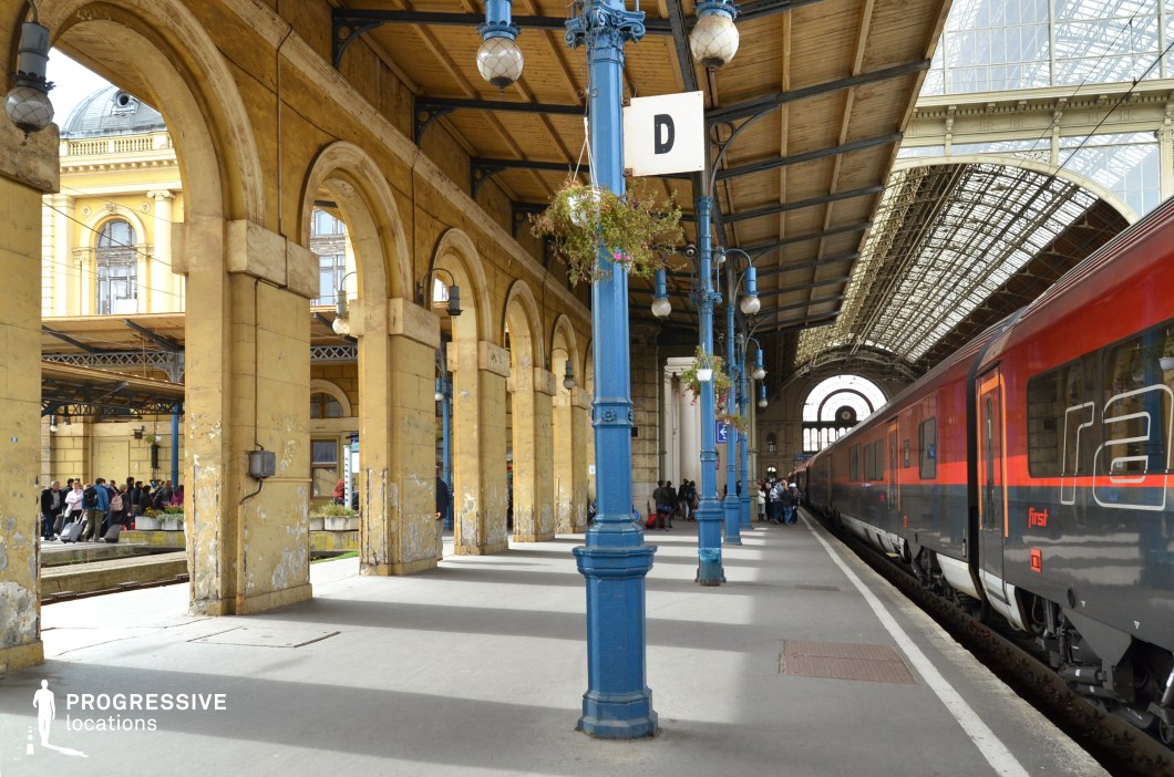 A grand train station platform with elegant arched columns and a towering glass-and-steel canopy, blending classic European architecture with industrial elements. The weathered yellow stone arches, adorned with intricate detailing, contrast beautifully with the deep blue cast-iron lamp posts that line the platform. The large glass windows allow natural light to illuminate the space, while a sleek modern train stands ready for departure, enhancing the juxtaposition of old and new. This visually rich and dynamic setting is ideal for filming travel sequences, dramatic farewells, or period scenes in genres such as historical dramas, thrillers, or romantic storytelling.