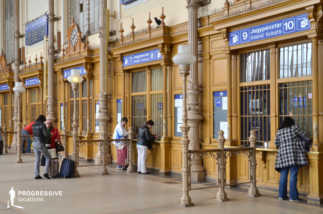 The image showcases a historic train station ticket hall with intricate architectural details. The wooden ticket booths are adorned with ornate carvings, framed by tall, fluted columns with decorative capitals. Above the booths, blue signage with white lettering indicates the ticket windows, blending seamlessly with the classic design. A vintage clock with an elaborate frame adds to the hall's nostalgic charm. The metal railings separating the waiting area from the ticket counters feature intricate wrought-iron patterns, enhancing the elegant atmosphere. The polished stone floor reflects the ambient lighting, complementing the warm, golden tones of the wooden structures. This location is ideal for filming period dramas, nostalgic travel scenes, or atmospheric mystery and noir films, offering a visually rich setting that evokes a sense of history and timeless elegance.