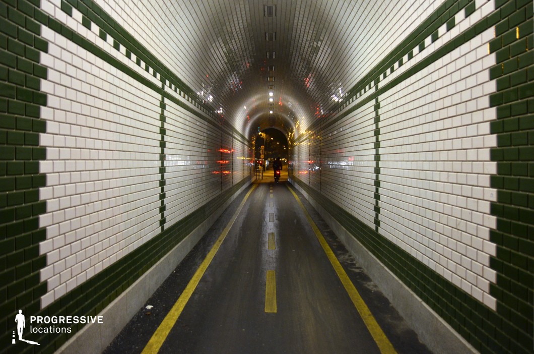 A narrow, arched tunnel lined with glossy white and green tiles creates a striking sense of depth and symmetry. The curved walls reflect ambient light and passing motion, producing dynamic distortions and subtle reflections. The dark asphalt pathway, marked with yellow lines, adds contrast to the structured geometric patterns of the walls. The distant exit reveals a cityscape illuminated by warm evening lights, enhancing the contrast between the confined tunnel and the open space beyond. This location is ideal for filming chase sequences, suspenseful moments, or psychological thrillers, offering a cinematic and immersive urban atmosphere.