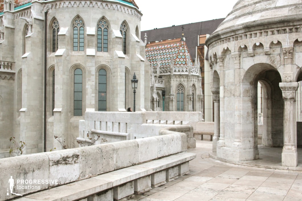 The image captures an architecturally rich location featuring intricate Gothic and Romanesque elements. The white limestone structures display pointed arch windows, decorative carvings, and a series of ornate columns and balustrades. A circular stone pavilion with arched openings and sculpted capitals adds depth to the scene, while the background features a grand church facade with a colorful tiled roof, evoking a medieval European aesthetic. The layered composition of staircases, terraces, and benches creates a visually dynamic environment. This setting is ideal for historical dramas, fantasy epics, and period-piece productions, offering an authentic and atmospheric backdrop reminiscent of medieval castles or sacred sites.