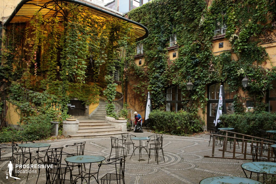 A secluded courtyard with a European charm, enclosed by ivy-covered walls that soften the aged yellow facade of the historic building. A curved staircase with ornate wrought-iron railings leads to an elevated entrance beneath a canopy draped in cascading greenery. The patterned stone pavement, complemented by vintage-style metal tables and chairs, enhances the old-world aesthetic. Large windows with dark frames and classic lanterns add depth and character to the space. This setting is ideal for filming scenes with a romantic, nostalgic, or mysterious tone, making it suitable for period dramas, artistic films, or European-style café scenes.