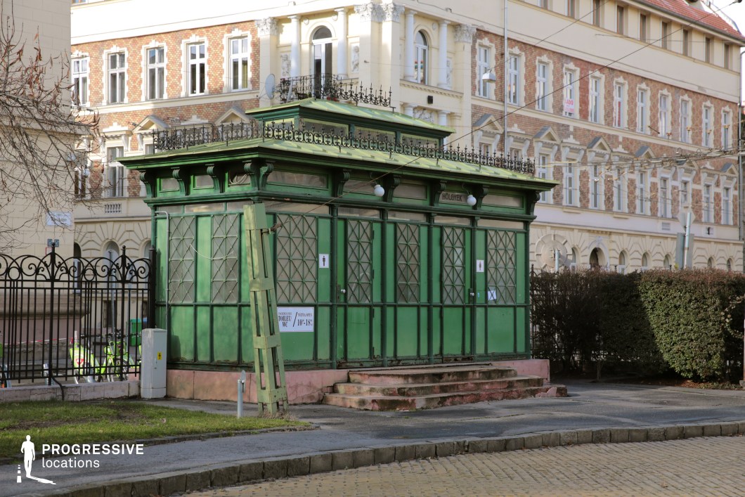 This image features a striking, historic green kiosk with intricate metal lattice windows and decorative wrought-iron detailing on its roof. The aged structure, possibly a public restroom or utility building, has a worn yet charming appearance, with peeling paint and a slightly leaning door adding to its character. Surrounding the kiosk is a cobblestone sidewalk and a backdrop of elegant historic buildings with red brick and cream-colored façades, adorned with arched windows and ornate cornices. The juxtaposition of the kiosk’s rustic appeal against the refined architecture makes this an excellent location for filming period dramas, urban documentaries, or atmospheric mystery scenes, offering a unique European city aesthetic.