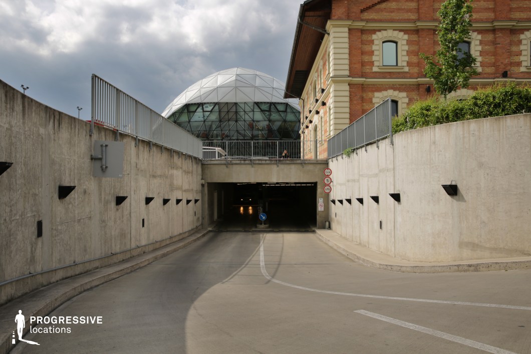 This image presents a striking contrast between architectural styles, featuring a curved concrete driveway leading into an underground parking facility. The stark, industrial aesthetic of the concrete walls is punctuated by angular black wall-mounted lights, creating a structured, repetitive visual pattern. Above the tunnel entrance, a historic brick building with arched windows and decorative stonework adds a classical touch, while in the background, a futuristic glass dome with a geodesic pattern introduces a modern element. The juxtaposition of old and new architecture enhances the location’s cinematic versatility, making it ideal for action sequences, high-stakes chase scenes, or dramatic thriller moments set in a contemporary urban environment.
