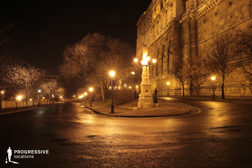 This elegantly curved road is lined with vintage street lamps casting a warm golden glow, complementing the towering historic stone walls of a grand building. The ornate lamppost at the central junction adds a regal touch, enhancing the old-world atmosphere. The reflective wet pavement and softly illuminated trees frame the scene, creating a moody and cinematic ambiance. This location is ideal for period dramas, mystery thrillers, or historical epics, offering a timeless and atmospheric European backdrop that evokes a sense of intrigue and grandeur.