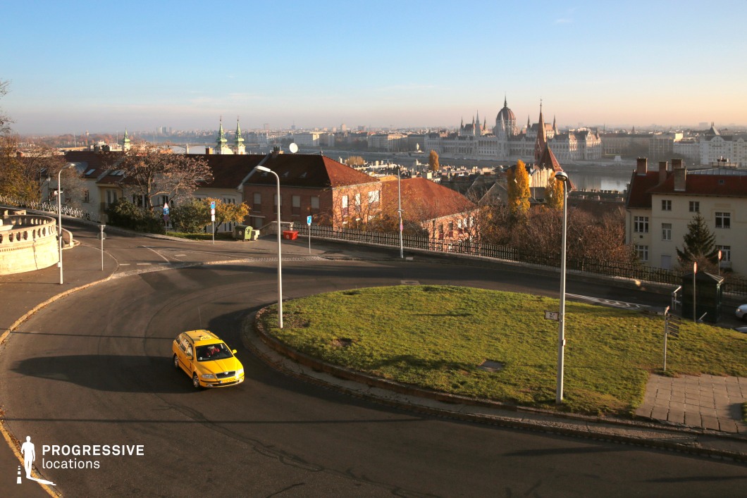 This elevated viewpoint captures a gently curving road encircling a small green space, surrounded by a mix of historic and modern architecture. The red-tiled rooftops and ornate facades contrast with the sleek, towering lampposts, creating a blend of old-world charm and contemporary urban infrastructure. In the background, an iconic neo-Gothic building dominates the cityscape, situated along a riverbank, enhancing the grand, cinematic depth of the composition. The curving road and varied elevation provide a dynamic setting ideal for dramatic driving sequences, establishing shots for urban narratives, or scenes depicting transitions between different city districts.