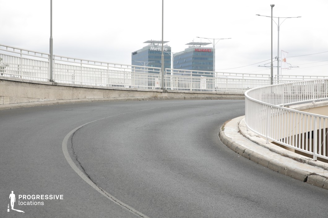 This modern overpass features a smooth, curving roadway lined with white metal railings, creating a clean and structured urban aesthetic. The upward angle of the road adds a sense of movement and anticipation, while the sleek, glass-covered high-rises in the background provide a contemporary corporate setting. The uniform lighting poles and minimalist infrastructure emphasize a functional, metropolitan design. This location is ideal for dynamic car chase sequences, futuristic cityscapes, or corporate thrillers, offering a sleek and modern backdrop with an emphasis on motion and urban architecture.
