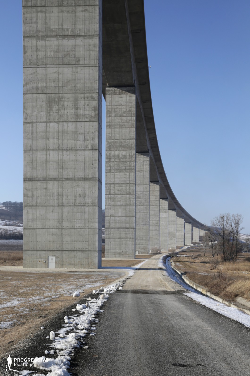 This image features an immense concrete viaduct stretching across an open landscape, supported by towering pillars that create a striking sense of scale and repetition. The raw, unadorned concrete surfaces give the structure an industrial and imposing aesthetic, emphasizing its sheer size and engineering precision. Below, a narrow road runs parallel to the viaduct, flanked by patches of dry grassland and a small drainage canal, enhancing the scene’s sense of isolation. The perspective lines lead into the distance, creating a strong visual depth. This location is ideal for dystopian or post-apocalyptic films, suspenseful road trip sequences, or dramatic establishing shots that emphasize solitude and vast infrastructure.