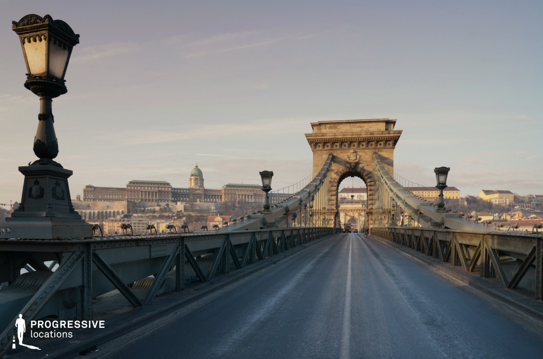 This striking urban scene features a historic suspension bridge with intricate ironwork and stone pillars, leading toward a grand neoclassical building complex in the background. The bridge's symmetrical design is enhanced by ornate lampposts, adding a sense of depth and perspective. The dominating structure in the distance, with its dome and extensive façade, provides a regal and monumental backdrop, reinforcing the European architectural grandeur. The muted color palette of stone, metal, and aged patina contributes to a timeless and cinematic quality. This location is ideal for period dramas, historical narratives, and dramatic urban sequences, offering a strong sense of place and architectural depth.