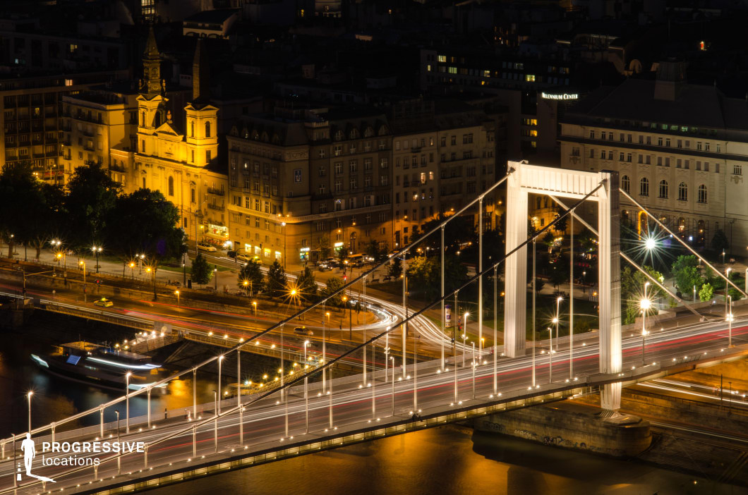 This striking night-time aerial view showcases a modern white suspension bridge stretching across a reflective river, with glowing light trails from moving vehicles enhancing the sense of urban dynamism. In the background, a warmly lit historic church and classic European buildings create a contrast between contemporary infrastructure and old-world architecture. The combination of vibrant city lights, intricate roadways, and the river’s shimmering surface makes this an ideal filming location for high-energy urban sequences, including action films, thrillers, or establishing shots that emphasize the grandeur of a European metropolis at night.