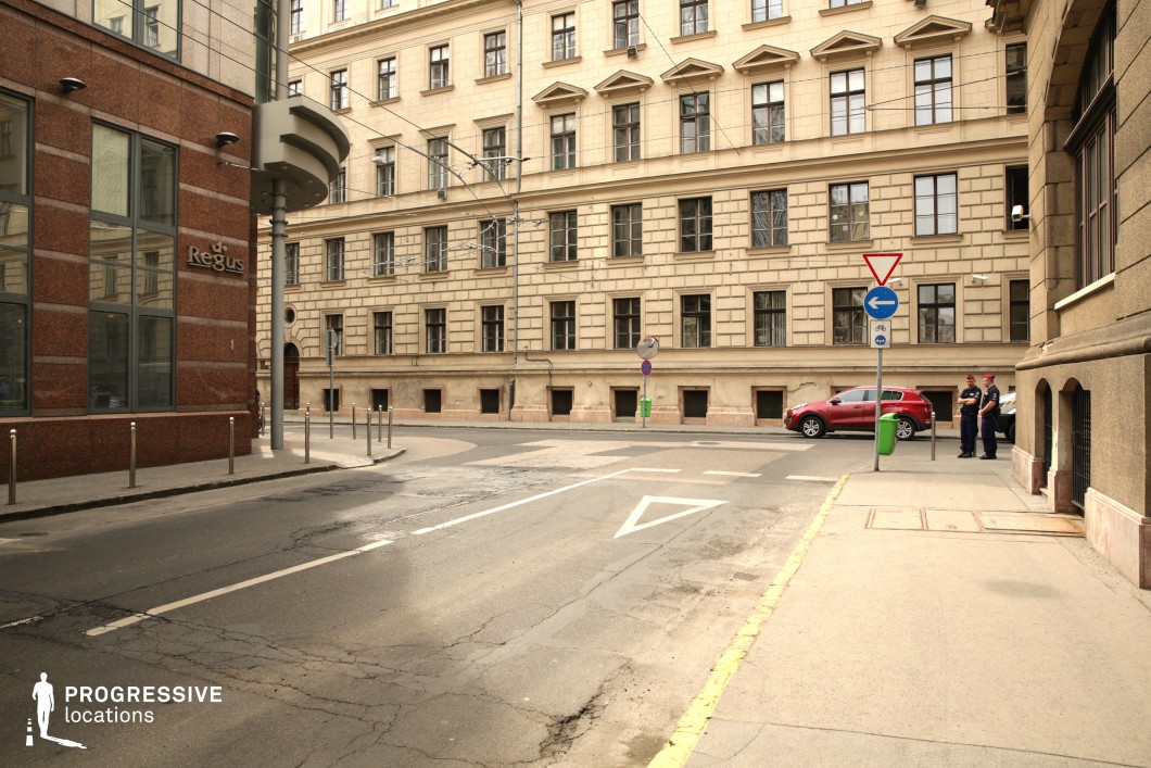 This European inner-city intersection features a blend of historic and modern architecture, with a beige neoclassical-style building dominating the background and a contrasting red granite-clad modern structure on the left. The street is narrow, with visible road markings, cracks in the asphalt, and subtle signs of wear, adding a sense of urban realism. Overhead tram cables reinforce the city’s functional infrastructure, while decorative window details and aged façades provide historical depth. This setting is ideal for urban drama, political thrillers, or investigative storylines, offering a balanced backdrop of institutional authority and everyday city life.