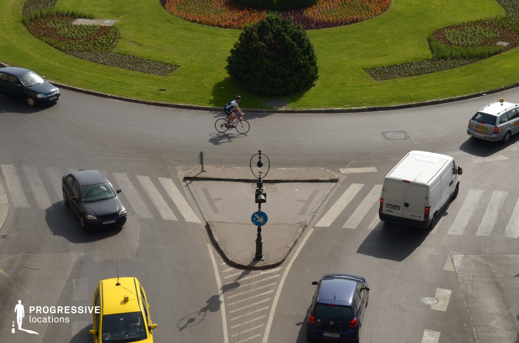 This top-down view of a triangular traffic island showcases an urban roundabout with carefully planned crosswalks, road markings, and a central lamppost adding a distinct focal point. Surrounding the intersection, a well-maintained green space with geometric flower arrangements creates a striking contrast to the asphalt and structured traffic flow. The image’s composition highlights symmetry and movement, making it an ideal setting for establishing shots, urban montage sequences, or dynamic chase scenes in dramas and action films.