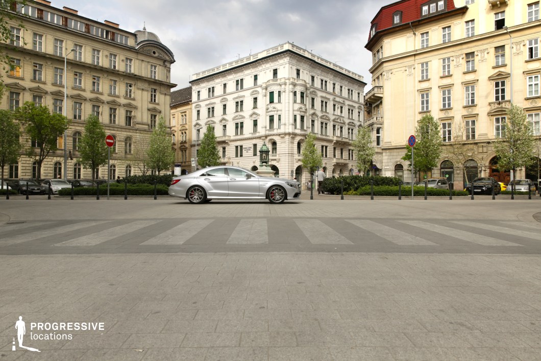 This refined urban square is surrounded by grand, historic buildings showcasing a mix of neoclassical and eclectic architectural styles, with ornate facades, arched windows, and decorative cornices. The symmetrical layout of the square is enhanced by neatly arranged trees, manicured greenery, and a pedestrian-friendly design marked by wide paving stones and a well-defined crosswalk. The combination of architectural grandeur and open space creates a sophisticated setting, ideal for high-end lifestyle scenes, period dramas, or establishing shots in films that require an upscale European city backdrop.
