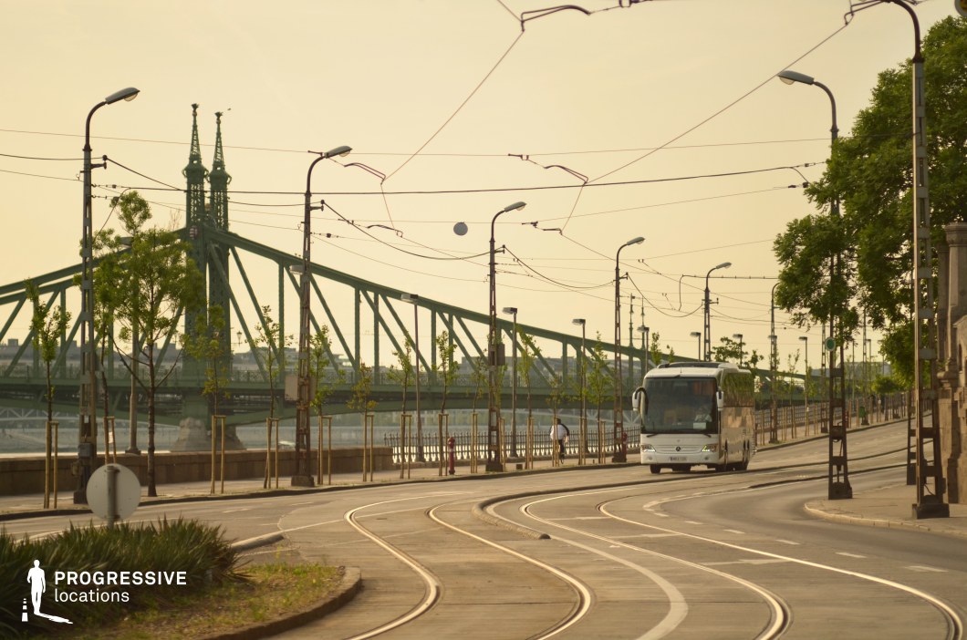 This urban riverside setting features a gently curving tramway lined with tall, vintage-style streetlights and overhead electric cables, creating a dynamic sense of movement. The striking green truss bridge in the background serves as a focal point, adding historical depth and architectural character to the scene. The metal railings, young trees, and neatly maintained greenery along the tram tracks contribute to a structured yet open atmosphere. This location is ideal for transportation-themed sequences, establishing shots of a European city, or narrative-driven scenes in dramas, thrillers, or romantic films that emphasize urban transit and daily life.