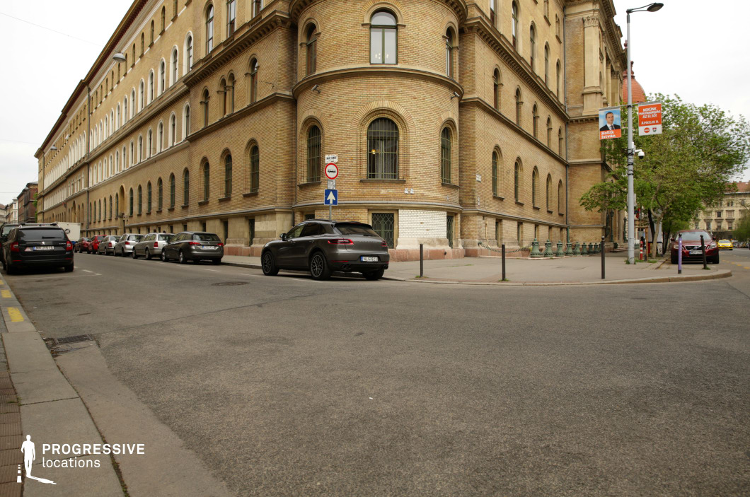 This urban street corner is dominated by a grand historic building with a rounded façade, featuring arched windows and warm-toned brickwork that exudes a sense of classical European architecture. The street layout, with parked cars lining the curb and a sidewalk framed by decorative metal poles, enhances the structured urban environment. The combination of the stately building and the slightly worn asphalt creates a balanced blend of elegance and realism. This location is well-suited for period dramas, mystery films, or crime thrillers, offering a timeless backdrop for both subtle character moments and high-stakes urban sequences.