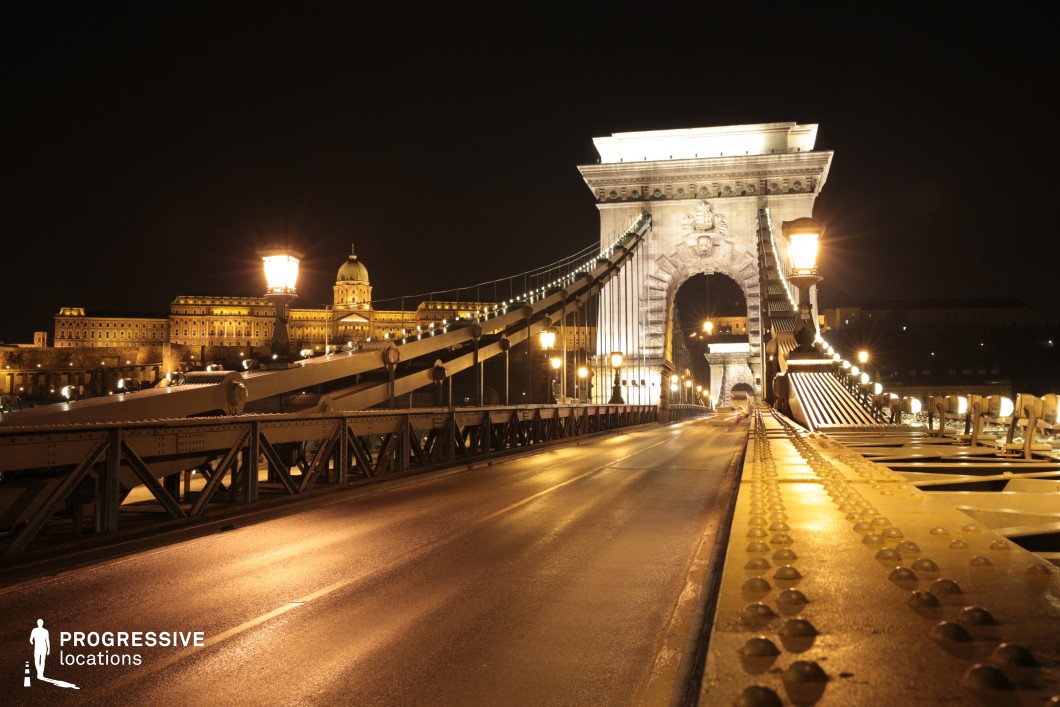 This grand suspension bridge, bathed in golden light, stretches across the scene with its massive stone gateway standing as an architectural centerpiece. The intricate iron framework and rows of glowing street lamps enhance the bridge’s stately presence, while the illuminated domed building in the background adds historical depth. The reflective wet road surface intensifies the cinematic quality, creating a dramatic contrast between light and shadow. This location is perfect for high-stakes thrillers, espionage dramas, or romantic nighttime sequences, offering a visually stunning and atmospheric urban setting.
