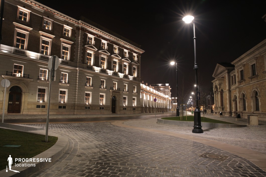 This grand, cobblestone-paved square is framed by stately neoclassical and baroque-inspired buildings, their facades highlighted by elegant architectural lighting. The warm glow of streetlights enhances the refined detailing of the structures, including arched windows, ornate cornices, and symmetrical proportions. A gently curving road and landscaped green spaces add to the balanced composition, while the polished surfaces of the pavement reflect ambient light, creating a cinematic night-time atmosphere. This location is ideal for period dramas, luxury brand commercials, or high-society intrigue, offering a refined and visually striking European setting.