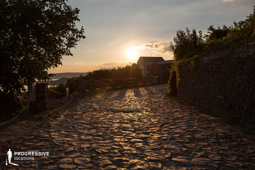 This image captures a winding cobblestone street bathed in warm, golden light, creating a dramatic interplay of textures and shadows. Flanked by a rugged stone wall on one side and a row of short stone pillars with rope fencing on the other, the path leads toward a small house nestled among lush greenery. The rough, uneven cobblestones contribute to the scene’s historic and rural charm, while the surrounding foliage and distant hills add depth. This location is ideal for period dramas, romantic narratives, or introspective character moments, offering a timeless and atmospheric setting rich in texture and warmth.