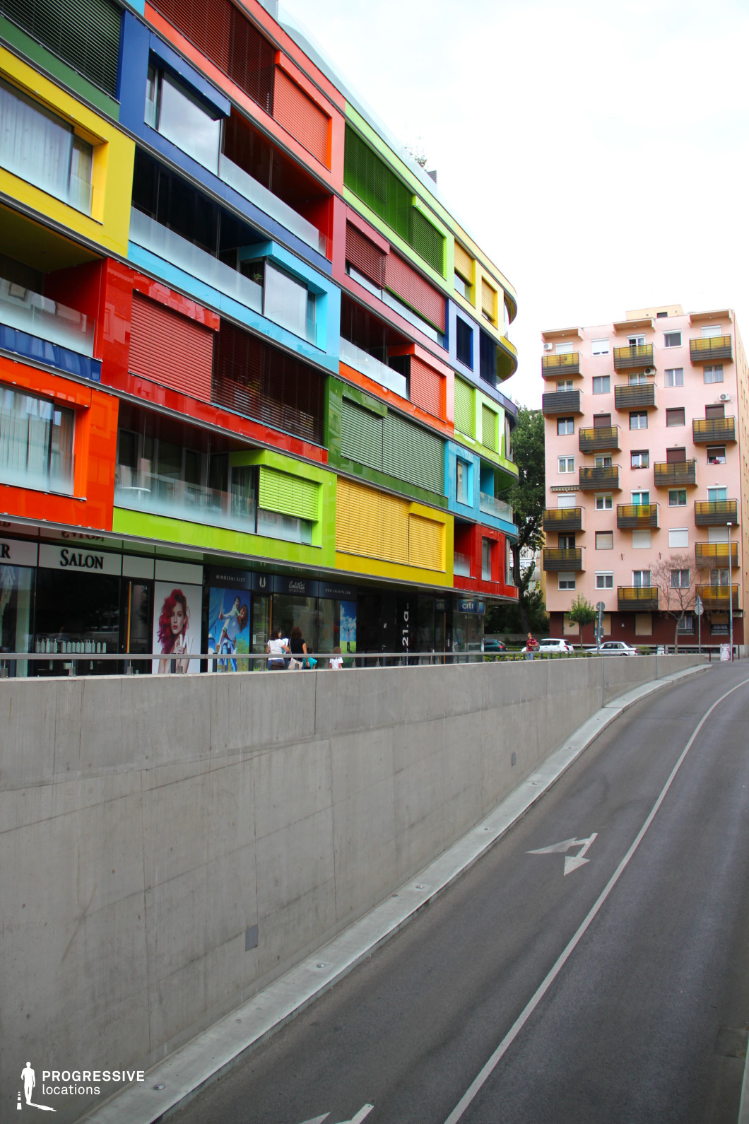 This image features a striking, multi-story modern building with a bold, colorful façade composed of geometric panels in red, yellow, green, and blue. The structure incorporates large glass windows, metal shutters, and sleek materials, giving it a contemporary and dynamic aesthetic. Below, a commercial level with storefronts interacts with the urban setting. A curving concrete road in the foreground leads upwards, bordered by tall retaining walls that create a sense of depth and movement. In contrast, an adjacent pastel-hued residential building with uniform balconies adds a subdued architectural element. The vivid colors and layered composition make this location ideal for fashion shoots, creative commercial productions, and films that require a vibrant, modern, and urban European atmosphere.