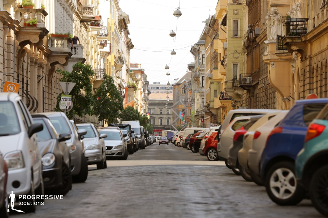 This image captures a narrow European street lined with ornately decorated historic buildings in warm tones of yellow, beige, and ochre. The façades feature intricate stone carvings, sculptural reliefs, and wrought-iron balconies, evoking a classical architectural style. Overhead, a string of suspended spherical streetlights enhances the depth and symmetry of the scene, drawing the eye toward the vanishing point in the distance. The cobblestone pavement adds a layer of texture, reinforcing the location’s old-world charm. This street is ideal for period films, romantic dramas, or mystery narratives that require a richly detailed European urban backdrop with a sense of timeless elegance.