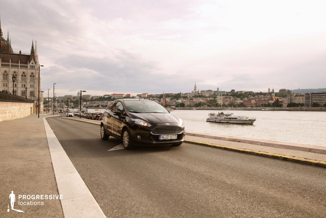 This gently curving riverside road runs alongside an iconic neo-Gothic building, with the calm waters of the river and a panoramic view of a historic cityscape in the background. The stone embankment creates a structured visual boundary between the roadway and the water, where boats navigate through the urban landscape. The perspective of the road, framed by lampposts and architectural details, enhances the depth and movement within the scene. This location is well-suited for establishing shots, chase sequences, or contemplative driving scenes in dramas, thrillers, and travel-based films.
