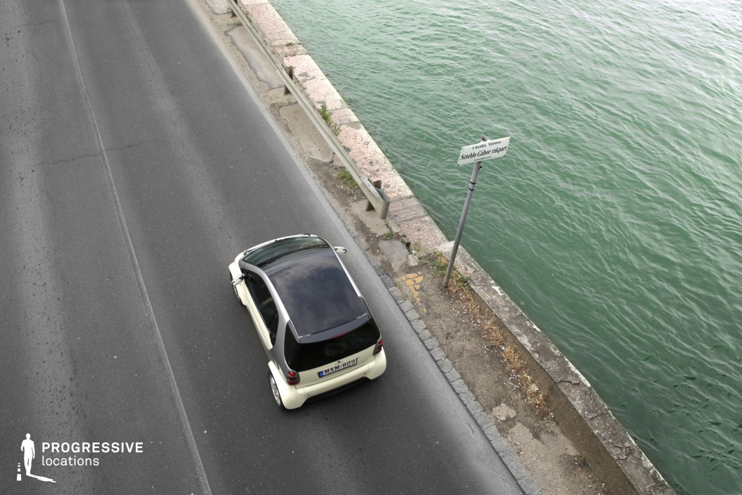 This aerial view captures a riverside road running parallel to a gently rippling waterway, separated by a weathered stone barrier. The slightly worn pavement, metal guardrail, and leaning street sign contribute to the urban realism of the scene. The muted color palette of asphalt, stone, and water enhances the location’s subdued yet atmospheric quality. This setting is ideal for introspective driving sequences, establishing shots in crime dramas, or contemplative urban narratives where the road and water serve as visual metaphors for movement and transition.